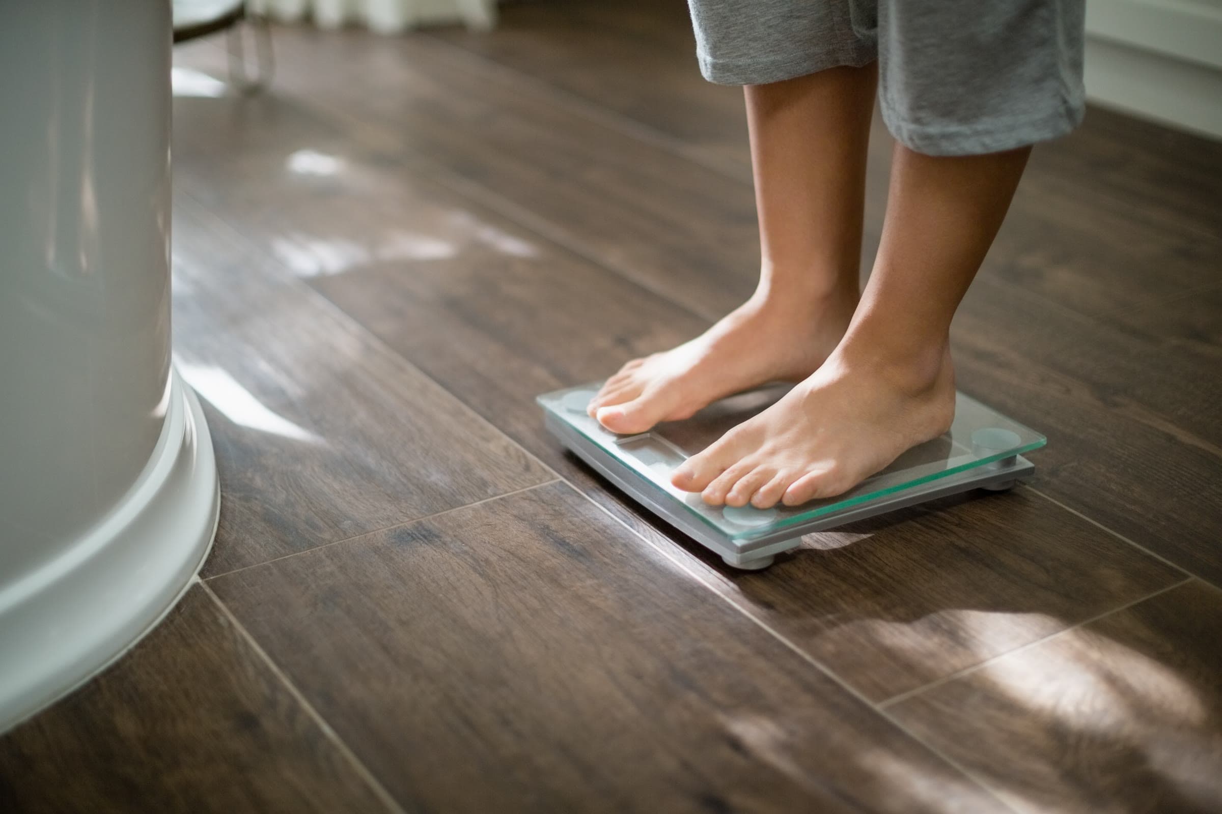 Bare feet in grey pyjamas standing on a glass bathroom scale on a wooden floor in morning light.